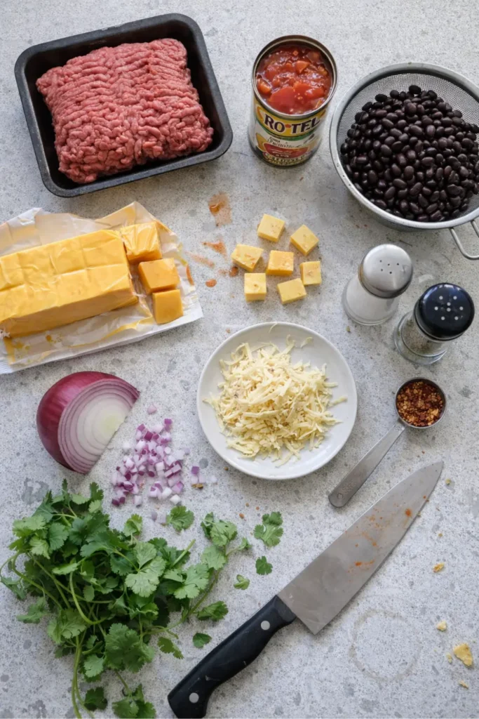 Flat lay of ground beef, Velveeta cubes, shredded pepper jack, Rotel, black beans, onion, cilantro, and spices on a gray countertop.